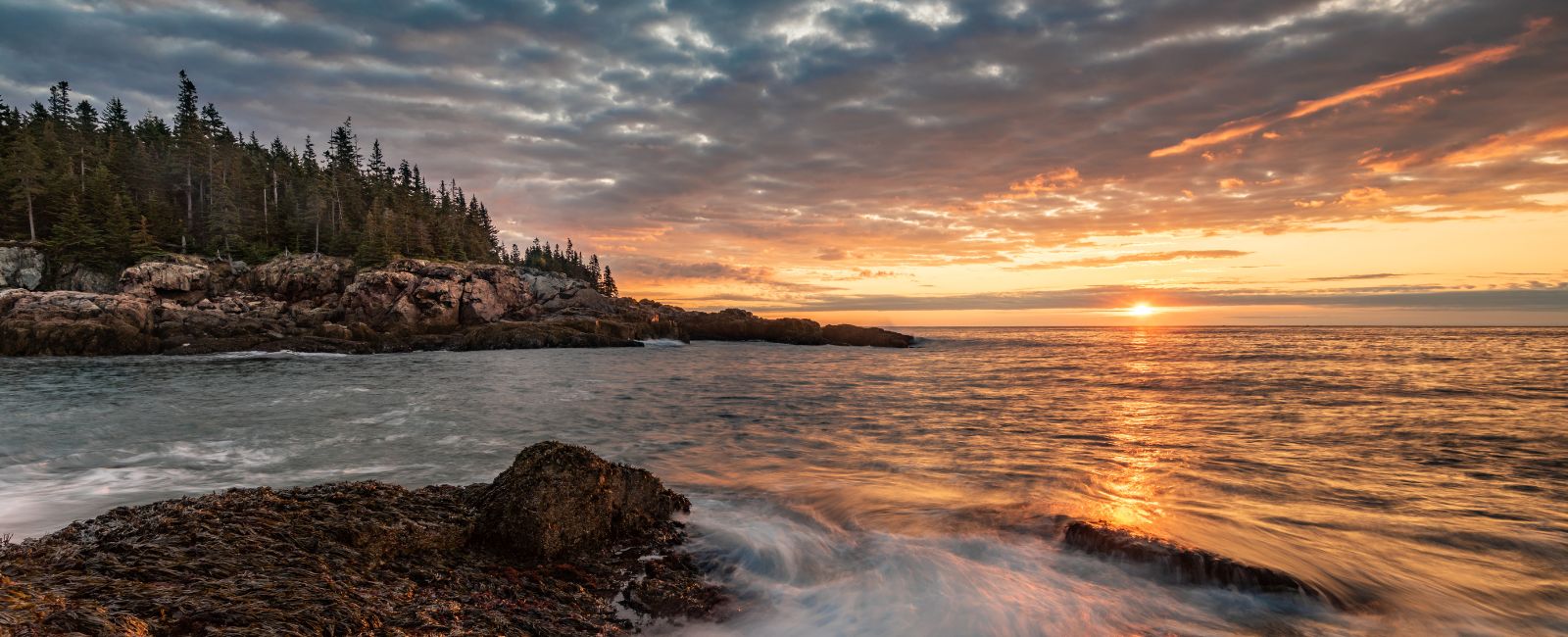 A cloudy sky and light orange sunset over the ocean and rocky shore at Acadia National Park in Maine