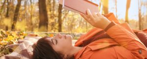 woman with brown hair wearing orange laying down on a blanket reading a book outside in autumn