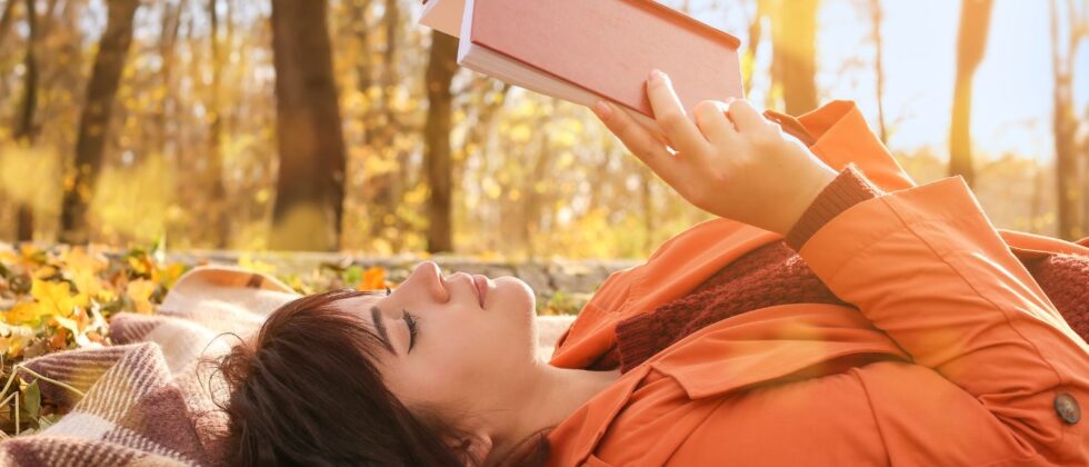 woman with brown hair wearing orange laying down on a blanket reading a book outside in autumn