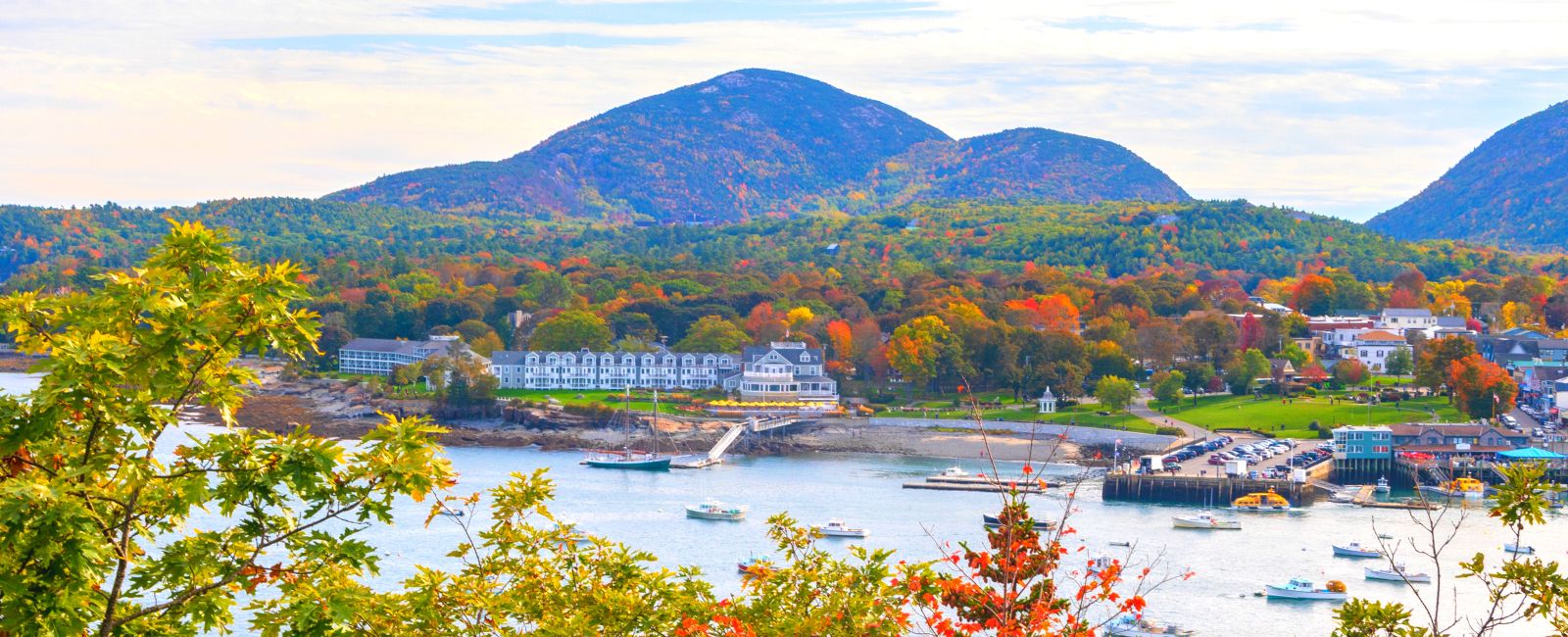 View of bar harbor maine from bar island, with trees in the foreground, then the ocean with boats, a large white building, a town off to the right, and autumn trees and mountains in the background
