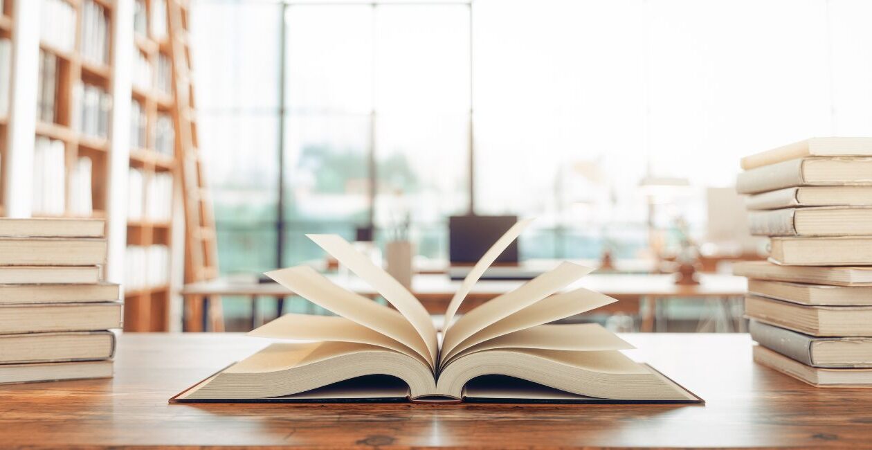 A library table filled with books and one in the center open with pages fanned in front of a blurred background of bookshelves and a wall of windows.