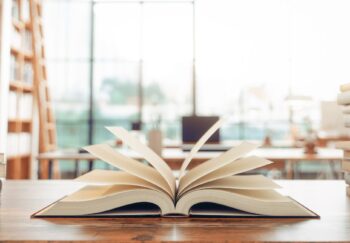 A library table filled with books and one in the center open with pages fanned in front of a blurred background of bookshelves and a wall of windows.