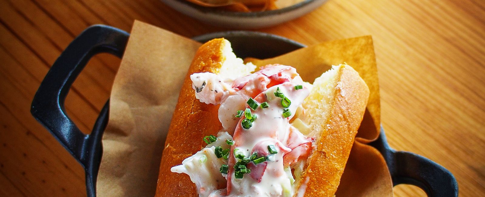 A close-up of a mini lobster roll with fresh, chopped chives in a pan sitting on a wooden table